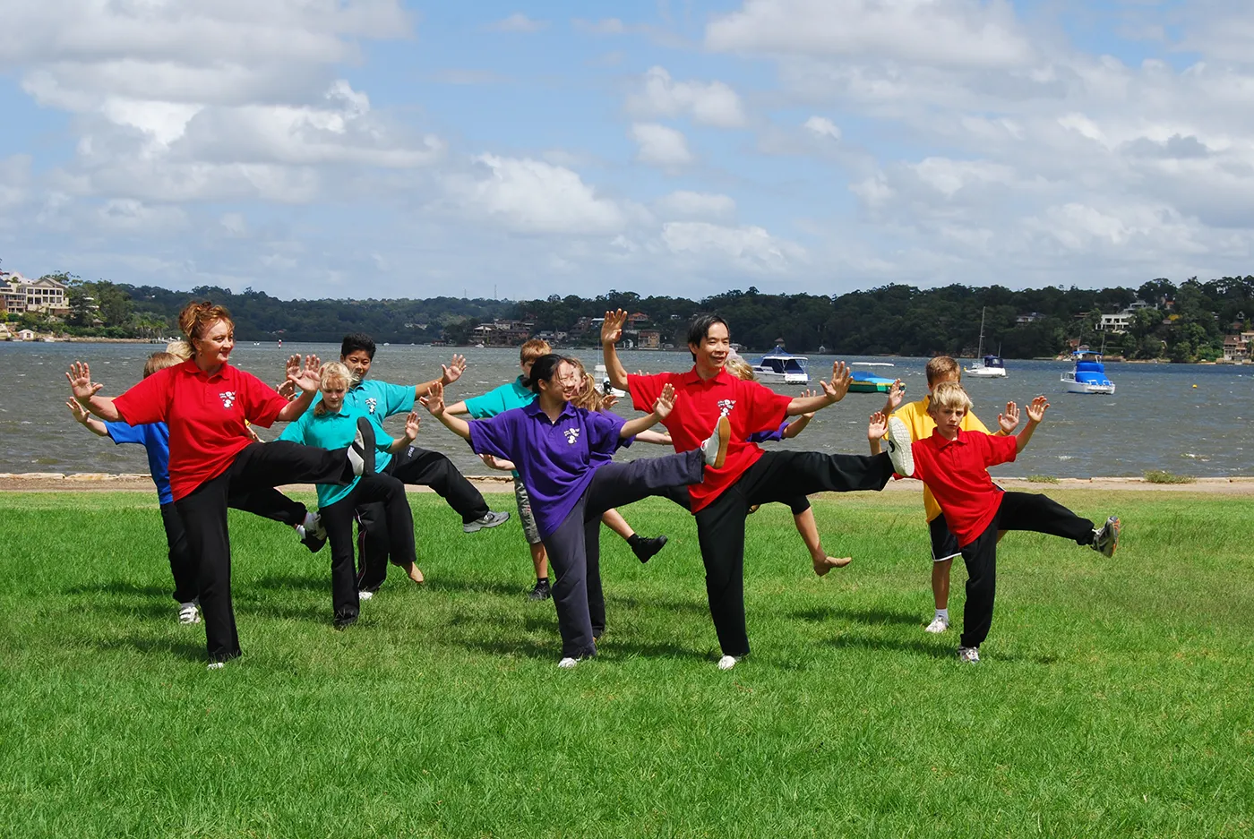 Dr Paul Lam and a group of children practising a tai chi kick on grass beside the water